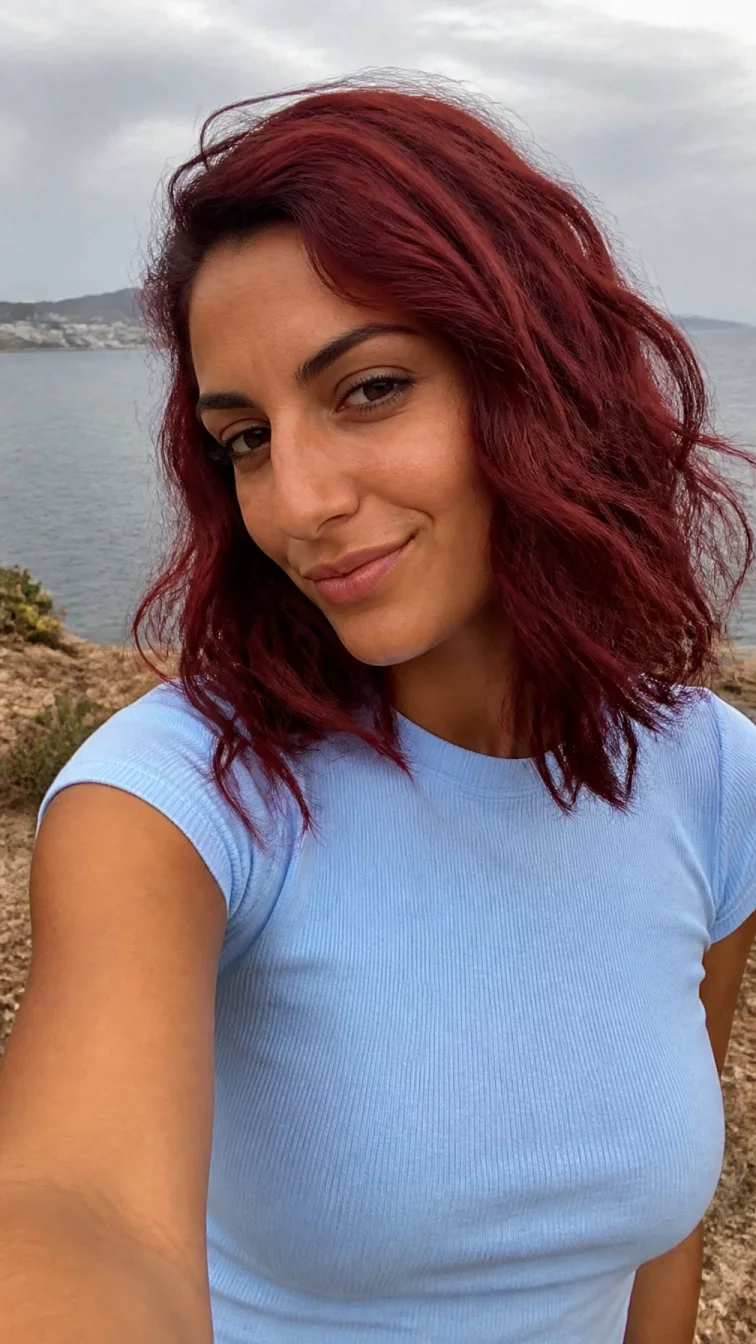 selfie of a 31-year-old Southern European woman with a square face, tan, aquiline nose, thin arched eyebrows, and full lips. Her face features slightly crooked smile, slightly flushed cheeks, and subtle natural facial asymmetry. She has downturned gray eyes, high cheekbones, subtle smile, playful. She is slim and is wearing a baby blue fitted t-shirt with spiced cider red wavy shoulder-length hair, overcast daylight, outdoor