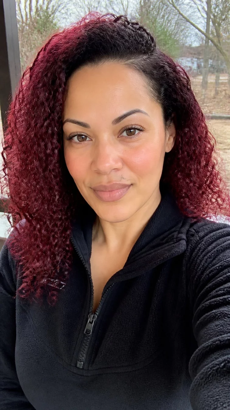 selfie of a 39-year-old Mixed-race woman with a diamond face, light, rounded nose, thin arched eyebrows, and wide lips. Her face features beauty mark near the lip, slightly flushed cheeks, and subtle natural facial asymmetry. She has almond-shaped brown eyes, pronounced cheekbones, neutral expression, cheerful. She is athletic and is wearing a black fleece pullover with deep burgundy wine curly hair, soft window light, outdoor