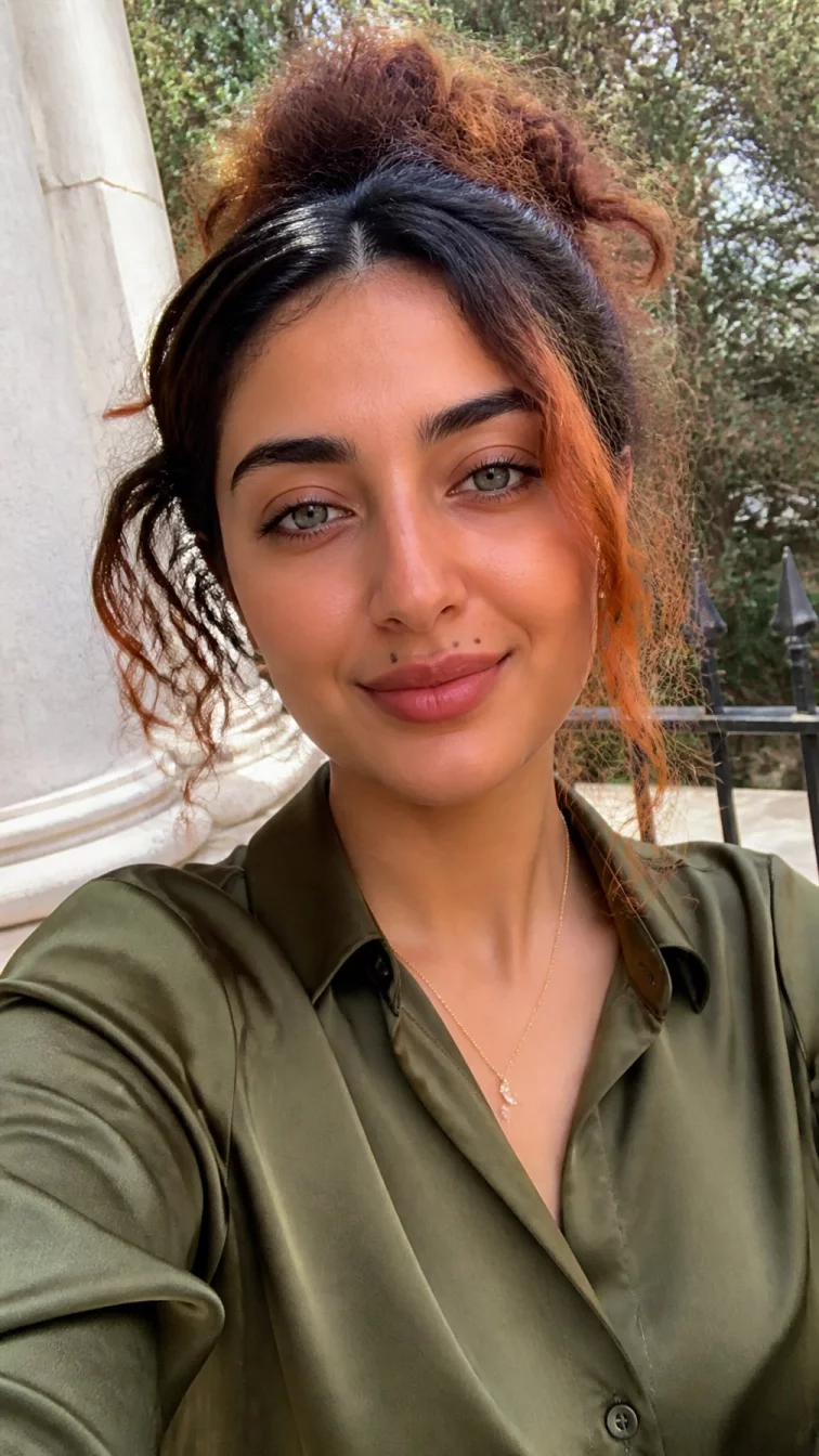 selfie of a 26-year-old Middle Eastern woman with a pear-shaped face, golden brown, aquiline nose, softly curved eyebrows, and full lips. Her face features beauty mark near the lip, subtle acne marks, and subtle natural facial asymmetry. She has hooded gray eyes, rounded cheeks, closed-mouth smile, thoughtful. She is slim and is wearing a olive green satin blouse with fiery burnt orange copper highlights messy bun hair, soft window light, outdoor
