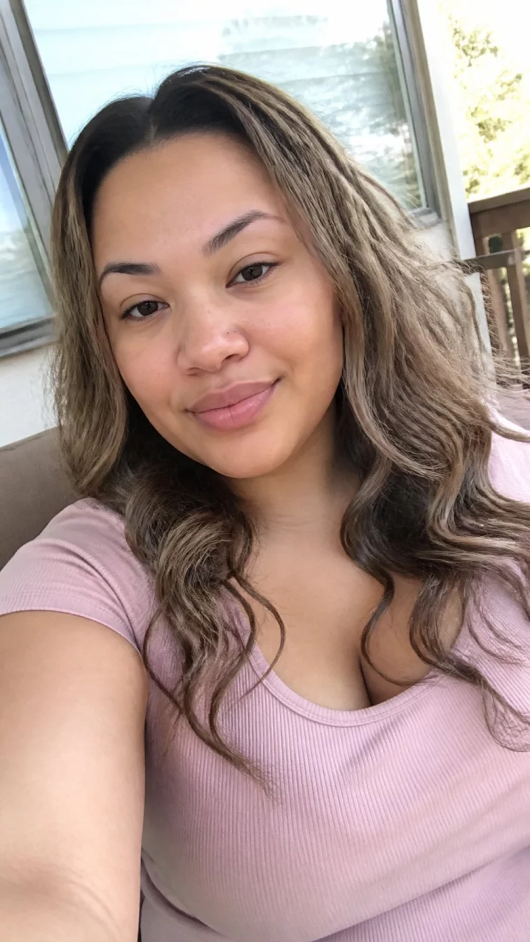 selfie of a 31-year-old Mixed-race woman with a round face, fair, narrow nose, softly curved eyebrows, and wide lips. Her face features widow's peak hairline, subtle acne marks, and subtle natural facial asymmetry. She has monolid brown eyes, soft cheekbones, closed-mouth smile, relaxed. She is curvy and is wearing a blush pink ribbed t-shirt with cool taupe chai highlights wavy hair, soft window light, outdoor