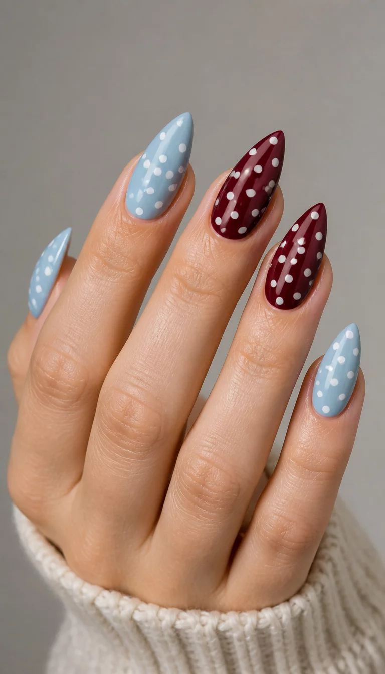 close-up shot of a woman’s hand with five fingers showing a mix of stiletto and almond-shaped nails, featuring light blue with white polka dots and burgundy with white polka dots, neutral studio background.