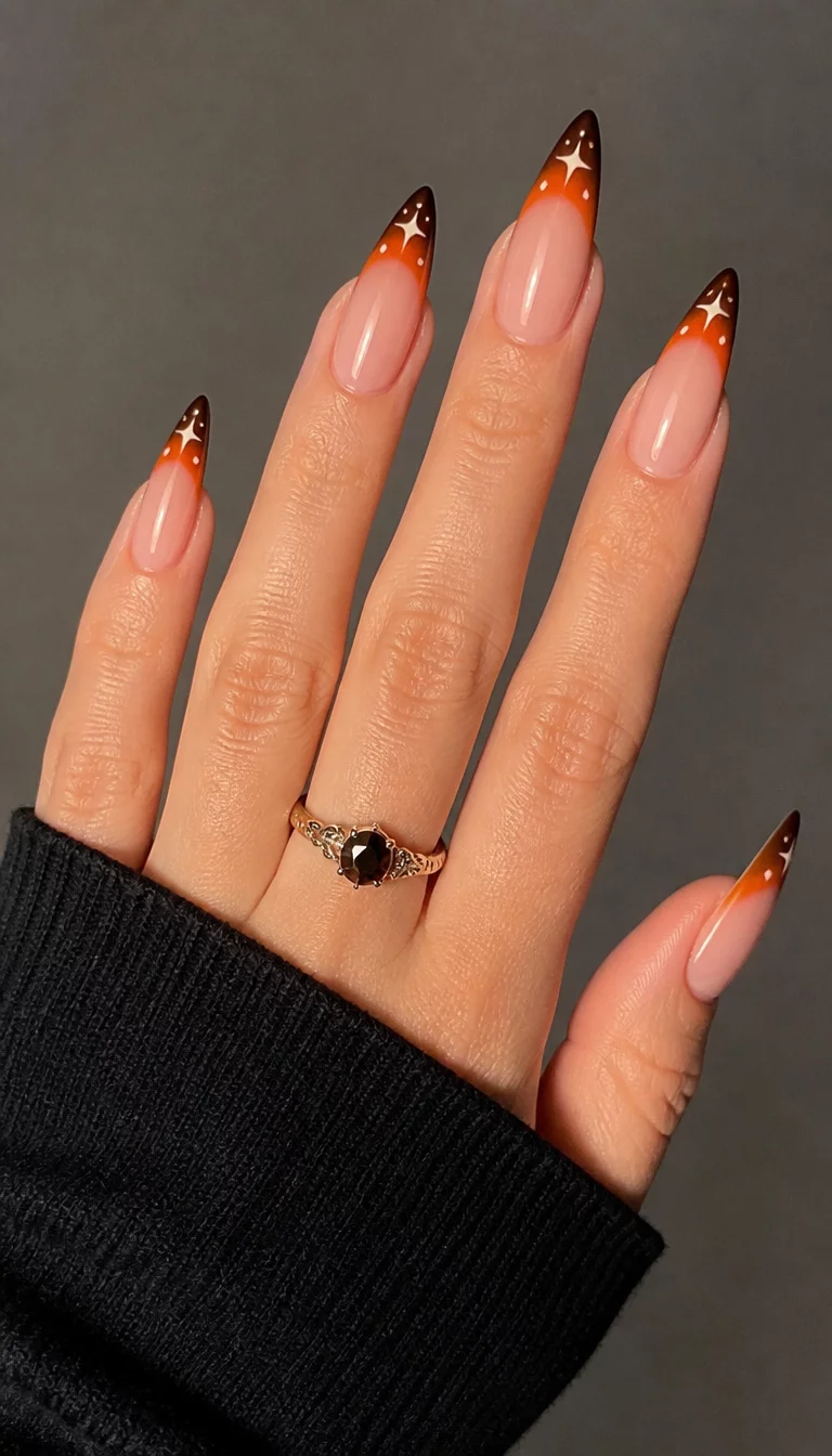 close-up shot of a woman’s hand with five fingers showing long pointed nails with a French tip featuring an ombre of orange and brown with small star designs, a black sleeve and a ring with a dark stone visible.