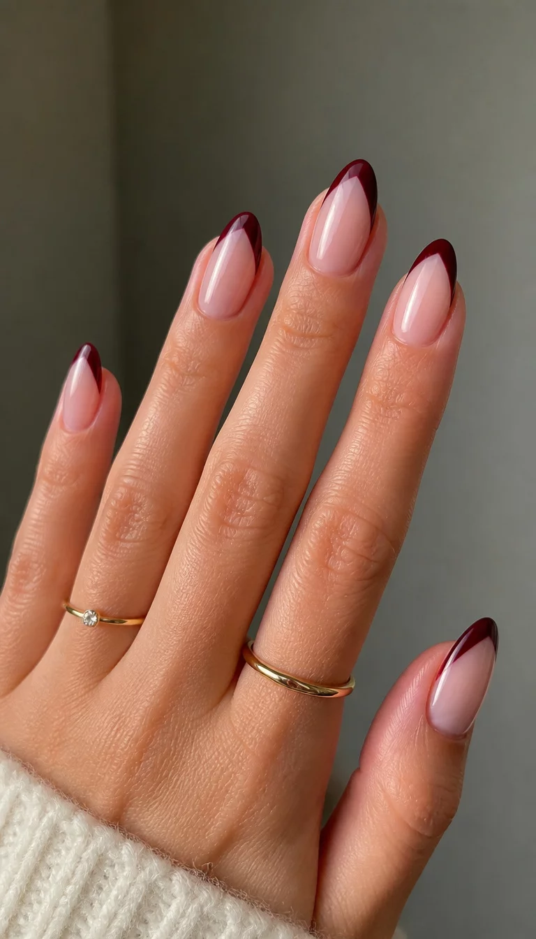 close-up shot of a woman’s hand with five fingers showing almond shaped nails with a sheer neutral base and a deep burgundy V-tip French manicure, two minimalist rings on the fingers.