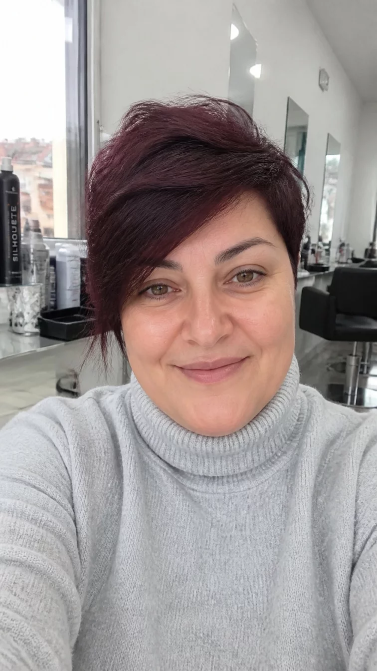 selfie of a 49-year-old Mediterranean woman with a round face, fair, narrow nose, thick natural eyebrows, and thin lips. Her face features uneven eyebrows, visible pores, and subtle natural facial asymmetry. She has upturned amber eyes, soft cheekbones, closed-mouth smile, relaxed. She is plus-size and is wearing a light gray turtleneck sweater with velvet burgundy asymmetrical pixie hair, overcast daylight, hair salon