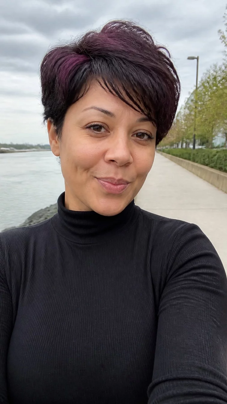 selfie of a 39-year-old Mixed-race woman with a pear-shaped face, light, rounded nose, thin arched eyebrows, and thin lips. Her face features dimples, subtle acne marks, and subtle natural facial asymmetry. She has almond-shaped dark brown eyes, pronounced cheekbones, closed-mouth smile, playful. She is curvy and is wearing a black mock neck sweater with blackberry swirl layered pixie hair, overcast daylight, outdoor