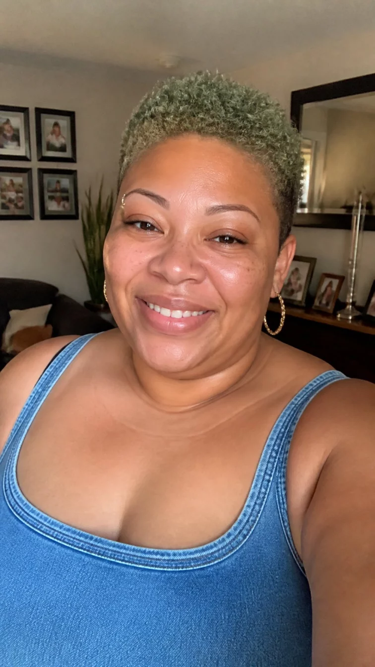 selfie of a 47-year-old Afro-Caribbean woman with a square face, light, broad nose, thin arched eyebrows, and wide lips. Her face features small scar on the eyebrow, light freckles, and subtle natural facial asymmetry. She has downturned amber eyes, high cheekbones, broad smile, relaxed. She is plus-size and is wearing a denim blue tank top with muted sage green curly pixie hair, warm indoor lighting, indoor