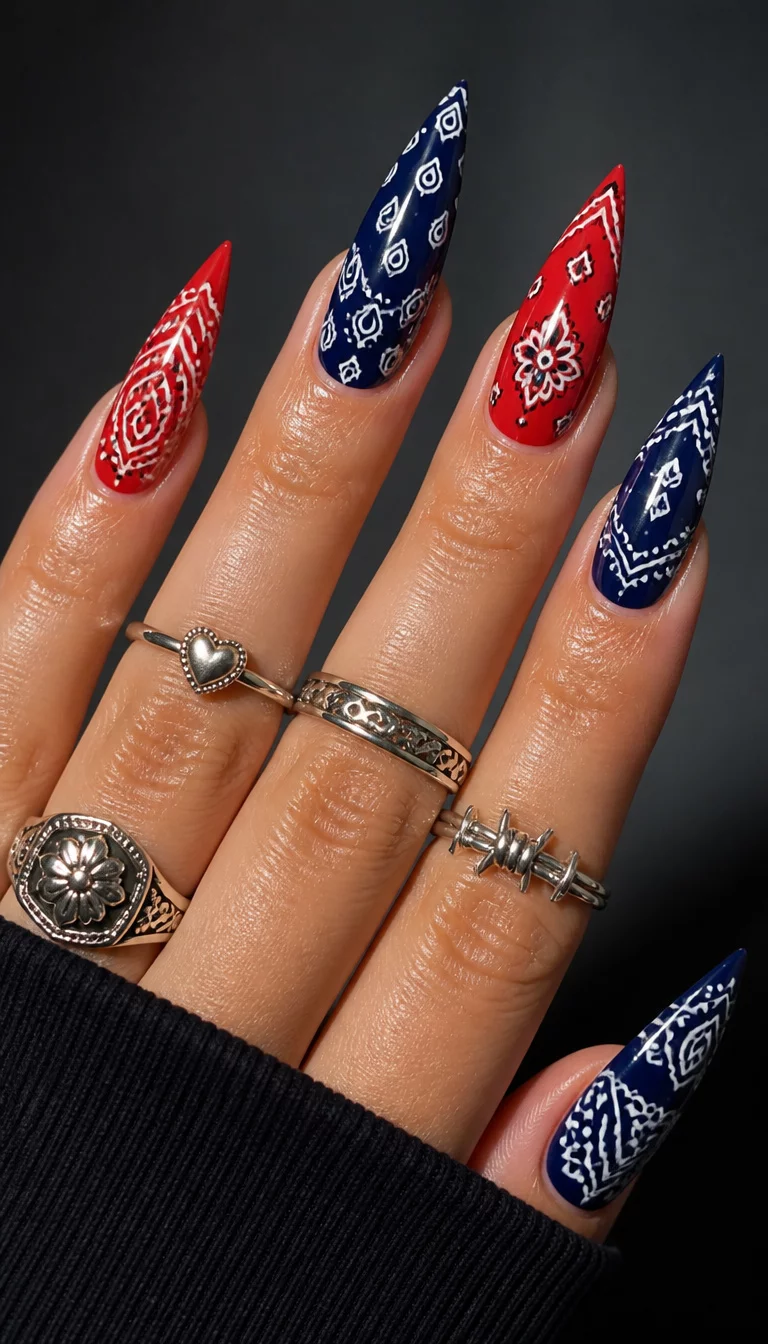 Midnight Bandana Claws close-up shot of a woman’s hand with five fingers showing stiletto nails featuring bandana patterns in red and dark blue, accented with white, adorned with several silver rings, indoor studio lighting.