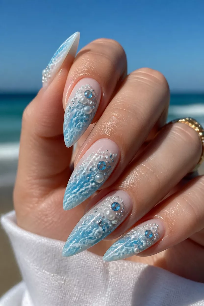 Ethereal Ocean Pearls close-up shot of a woman’s hand with five fingers showing nails with long sharp almond nails with light blue and white swirls, white pearls, and blue crystal gems, mimicking sea foam.