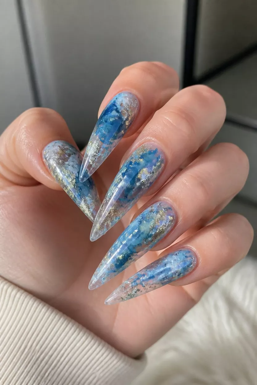 Dark Foil Drama close-up shot of a woman’s hand with five fingers showing nails with very long stiletto-shaped artificial nails with a blue, white, and translucent smoky base accented with gold and silver foil, indoors.