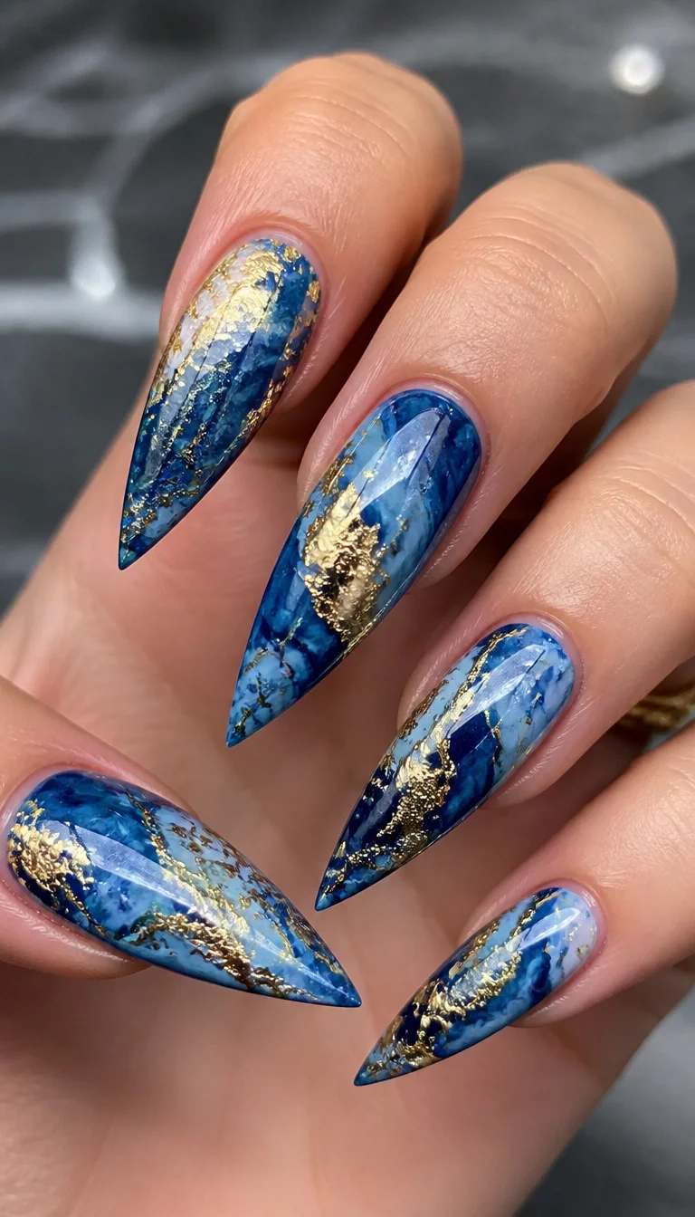 Marble Blue Gold Stilettos close-up shot of a woman’s hand with five fingers showing nails with long pointed stiletto nails with blue and gold abstract marble design, crushed foil effect.