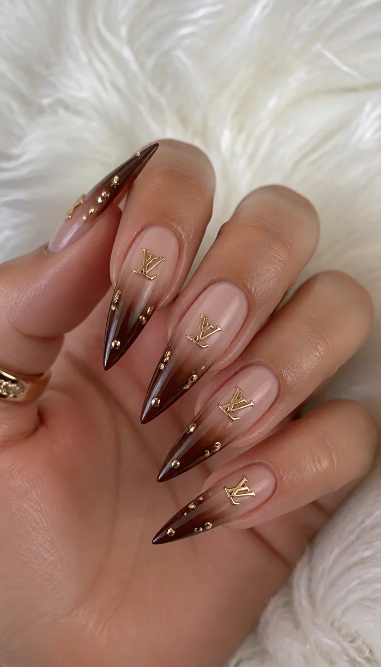 close-up shot of a woman’s hand with five fingers showing long, stiletto-shaped nails transitioning from a neutral base to a deep brown tip, adorned with gold logos and small gold studs, fluffy white background