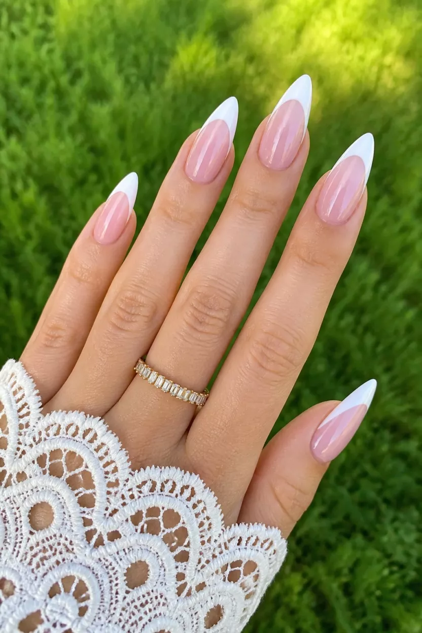 close-up shot of a woman’s hand with five fingers showing nails with long, medium-stiletto nails featuring a sheer pink base with classic white French tips, glossy finish, outdoors with green grass and white lace visible.