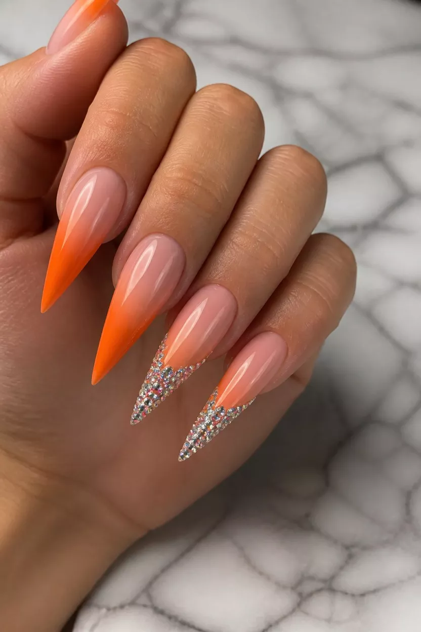 close-up shot of a woman’s hand with five fingers showing nails with very long, sharp stiletto nails painted in a vibrant neon orange ombre, two fingers heavily encrusted in small silver rhinestones along the free edge, marble surface background.