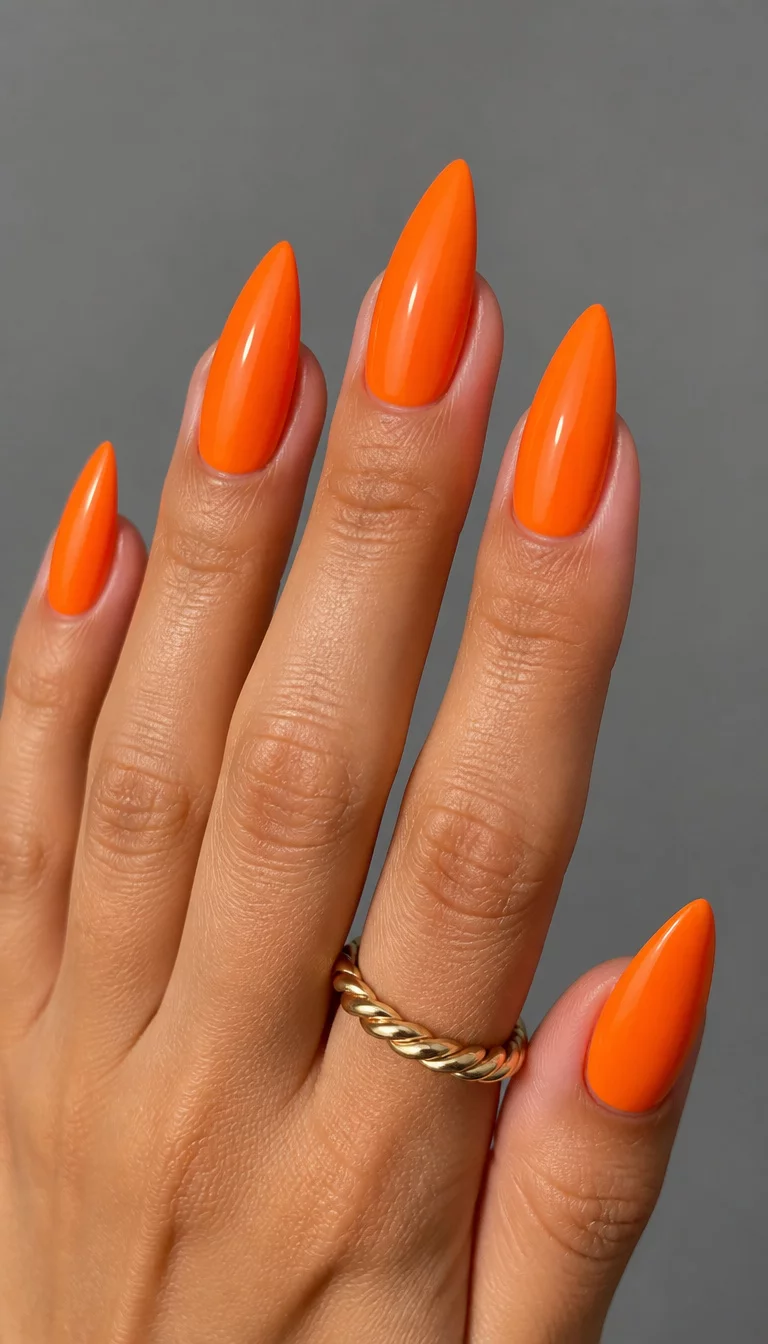 Sleek Stiletto Energy close-up shot of a woman’s hand with five fingers showing nails with stiletto shaped nails painted in a bright solid orange polish, with a simple gray background and a twisted gold ring.