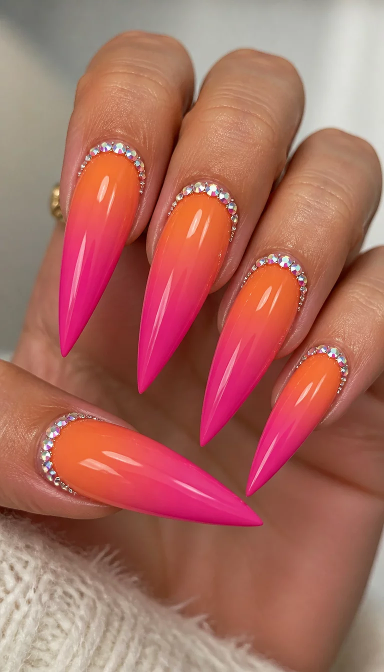 Rhinestone Stiletto Sunset close-up shot of a woman’s hand with five fingers showing nails with long sharp stiletto style nails with an orange to pink ombre effect and adorned with rhinestone accents along the cuticle.