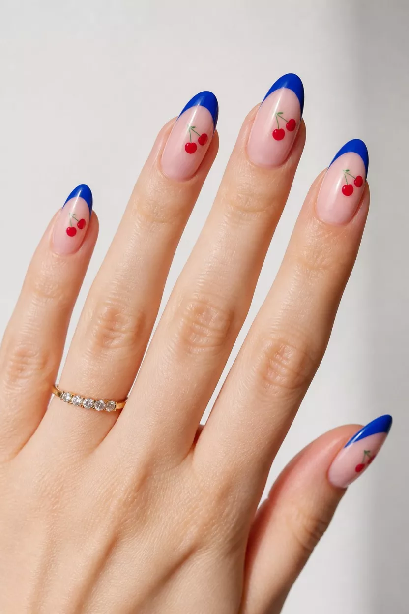 close-up shot of a woman’s hand with five fingers showing nails with almond-shaped nails with a light pink base and bright blue French tips and small red cherries, light background.