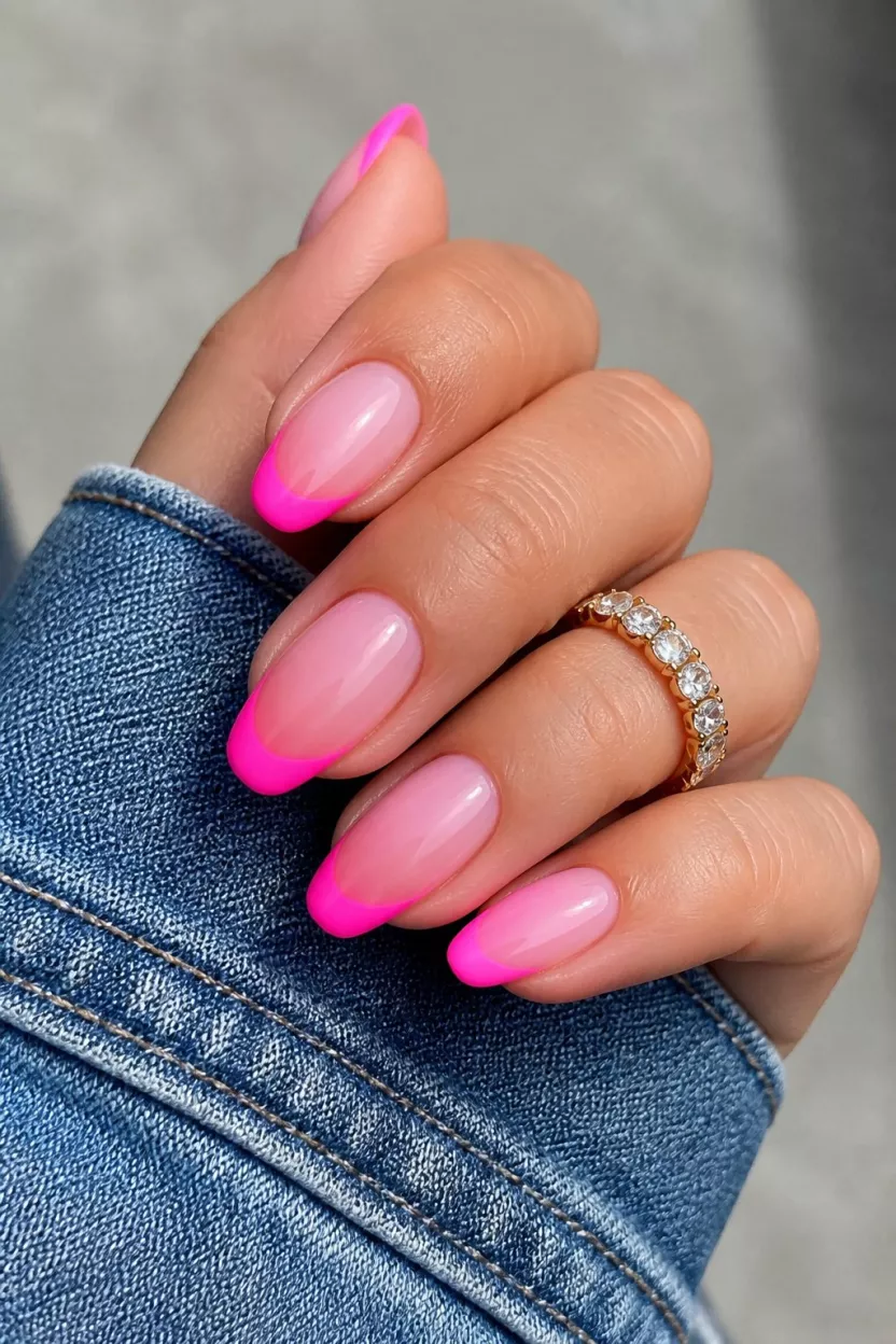 close-up shot of a woman’s hand with five fingers showing nails with medium length almond nails featuring a pink ombre base and neon hot pink French tips, denim sleeve background.