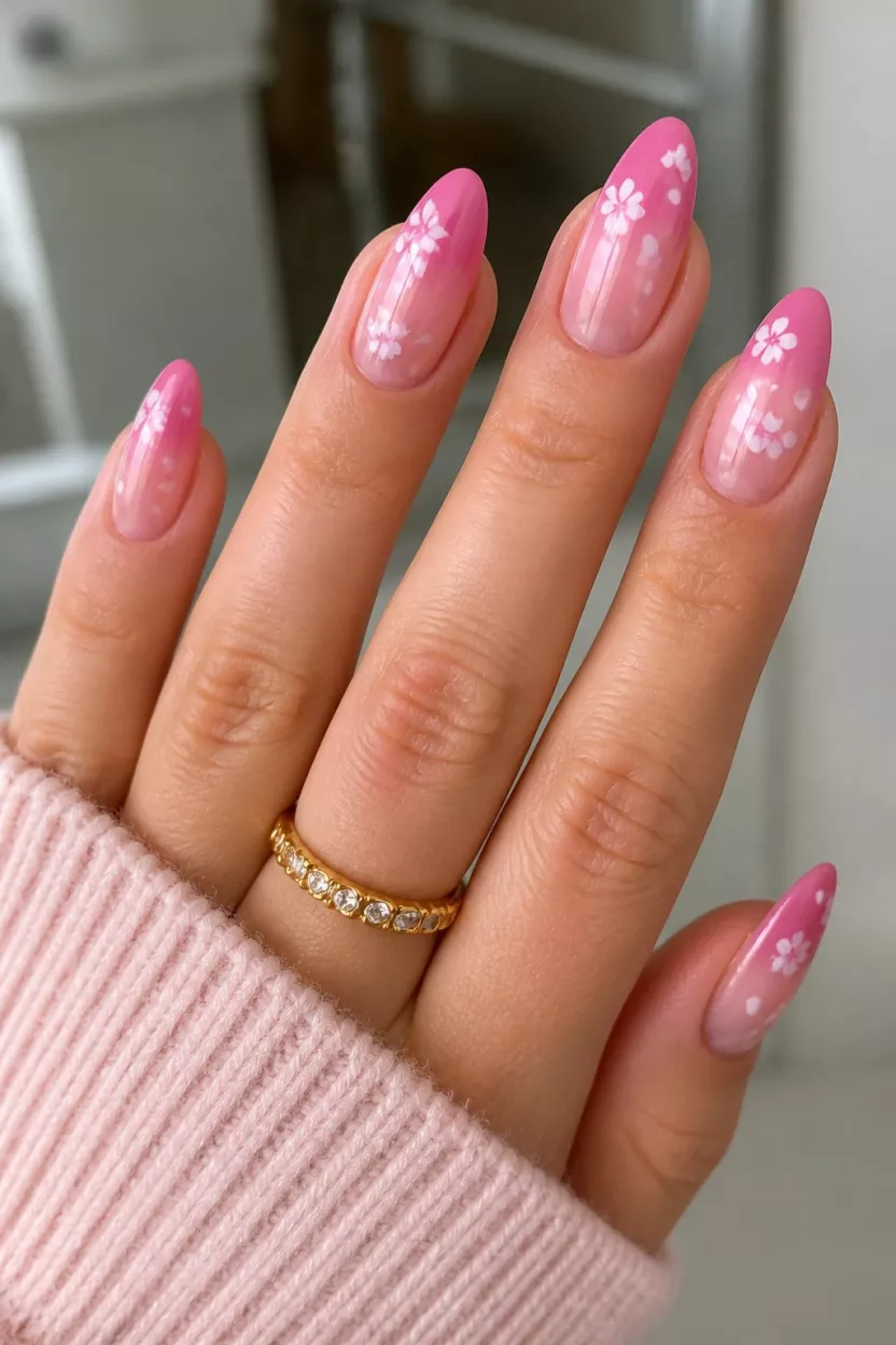 close-up shot of a woman’s hand with five fingers showing nails with medium-long almond nails featuring a pink ombre fading from deep pink to sheer with dainty white flowers