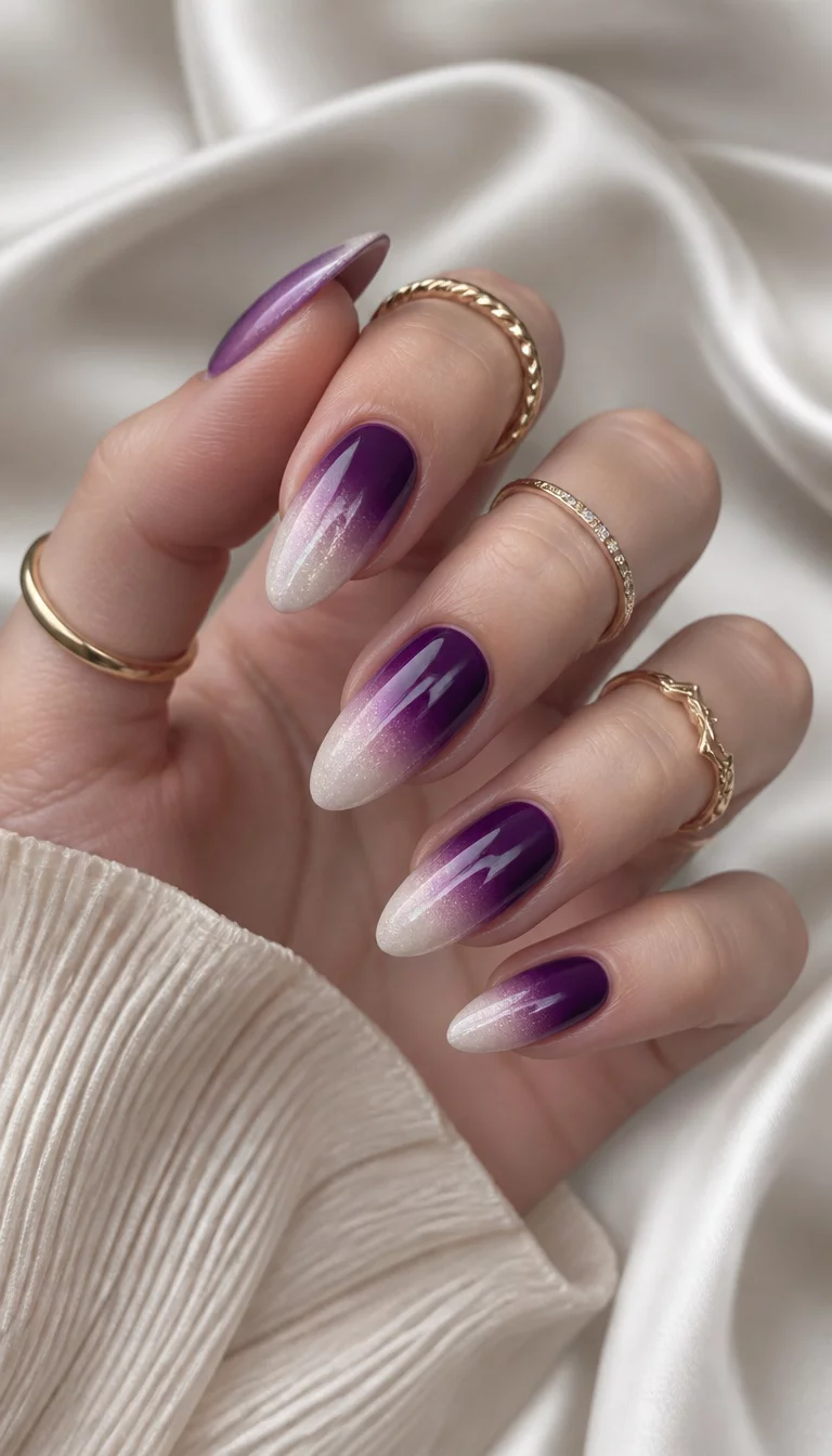 Deep Gradient And Gold Rings close-up shot of a woman’s hand with five fingers showing nails with almond-shaped nails featuring a gradient of deep purple fading into white gold rings on fingers, soft draped fabric background.