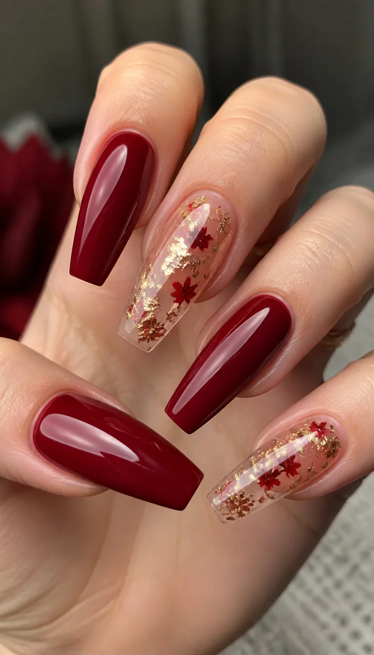 close-up shot of a woman’s hand with five fingers showing long coffin-shaped nails alternating between solid deep red and clear nails with gold leaf and red leaf confetti