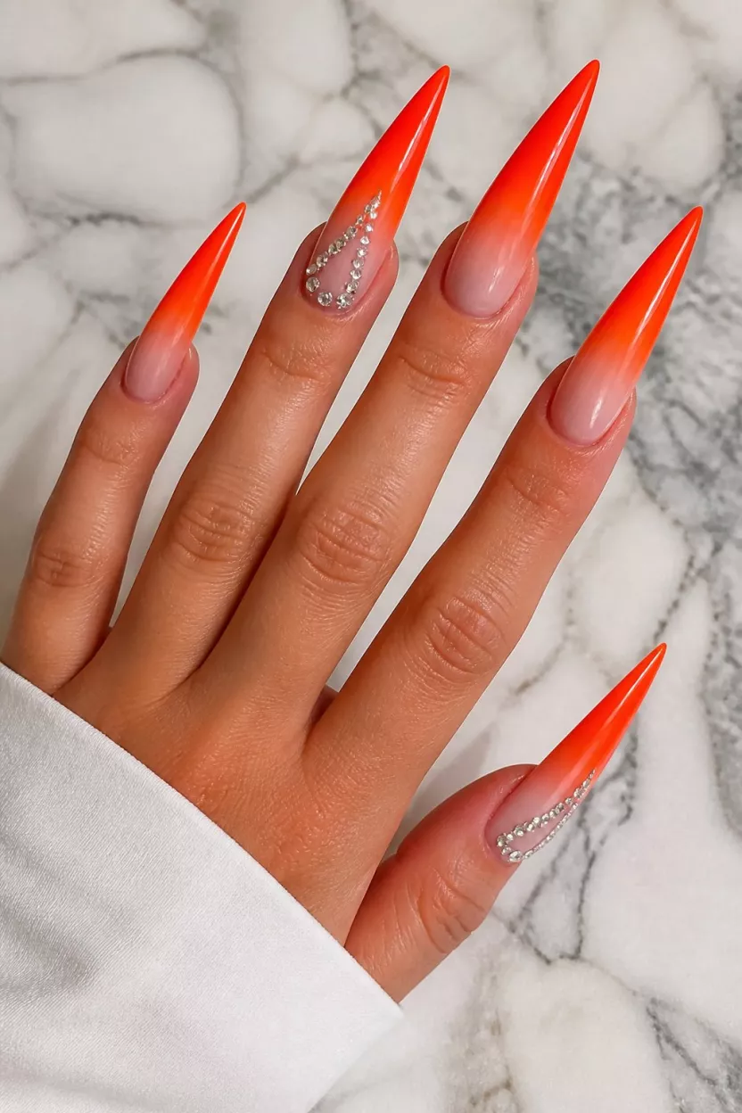 close-up shot of a woman’s hand with five fingers showing nails with very long stiletto-shaped nails featuring a bright orange-red ombré fade, accent nails with lines of small silver rhinestones, over a marbled surface.