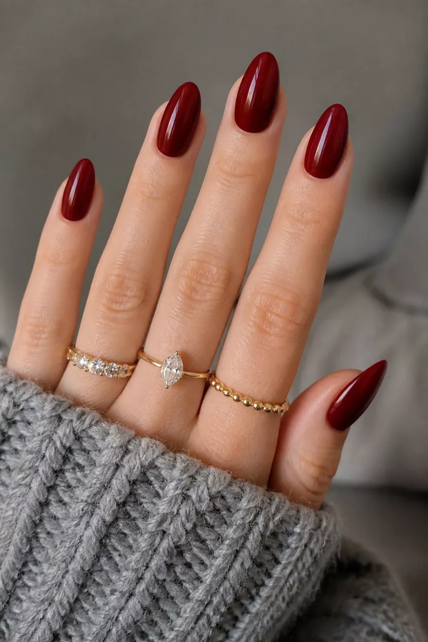 close-up shot of a woman’s hand with five fingers showing nails with long almond-shaped nails in deep glossy ruby red, gold rings, textured grey sweater.