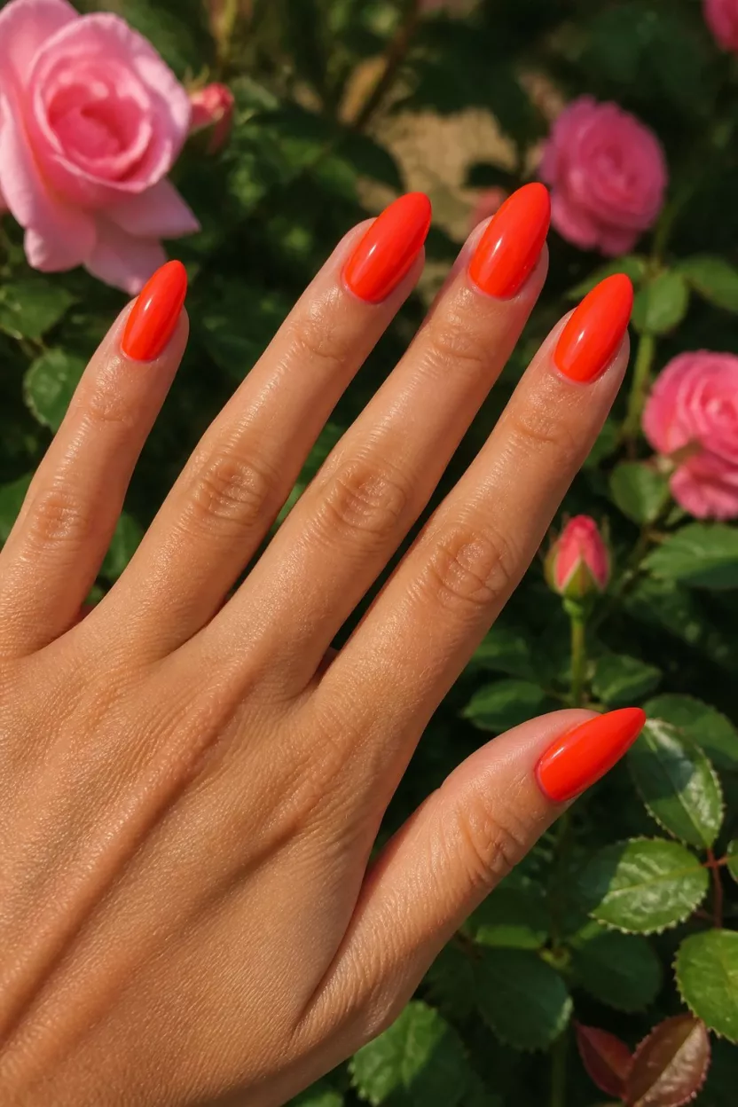 close-up shot of a woman’s hand with five fingers showing nails with long almond-shaped nails in neon fiery orange-red, outdoors with green foliage and pink roses.