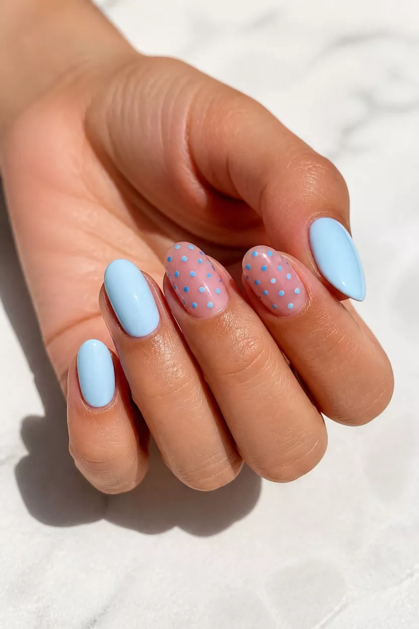 Pale Blue Polka Dots close-up shot of a woman’s hand with five fingers showing oval nails painted a pale pastel blue, with some featuring small, bright blue polka dots over a sheer base, against a bright, shadowed white surface.