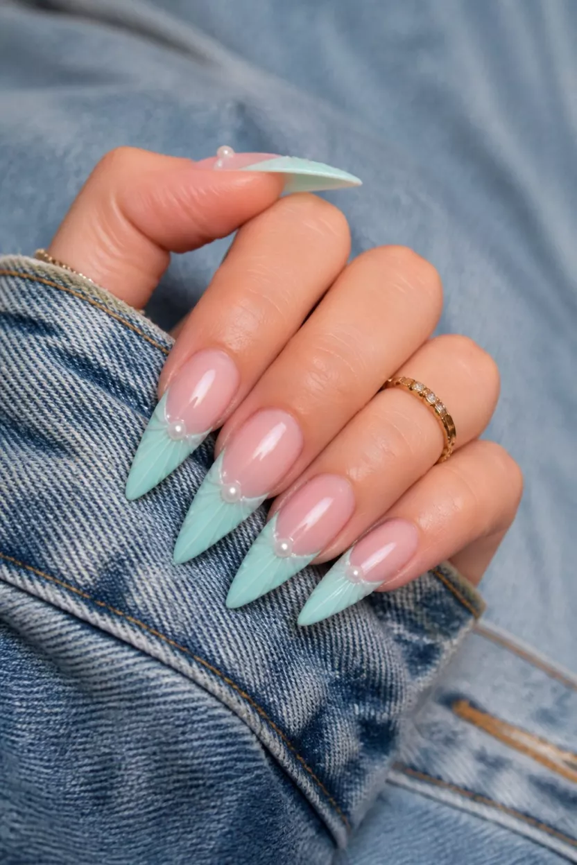 Mint Green Seashell Stilettos close-up shot of a woman’s hand with five fingers showing long, sharp stiletto nails with a sheer base and a mint green, scalloped French tip design resembling seashells, accented by small pearls, over a denim background.