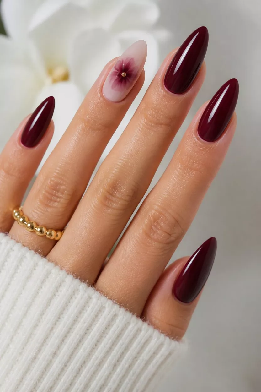 Rich Burgundy And Nude Florals close-up shot of a woman’s hand with five fingers showing almond and stiletto nails with a rich, deep burgundy polish and a nude base accent nail featuring a diffused burgundy flower design with a gold stud center.
