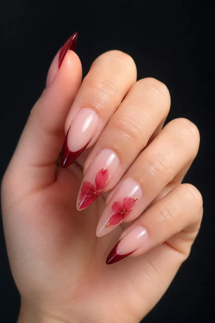 Deep Red French Tips With Botanical Details close-up shot of a woman’s hand with five fingers showing pointed stiletto nails featuring a deep red French tip with a diffused red flower design on accent nails, accented by gold studs, set against a dark, neutral background.
