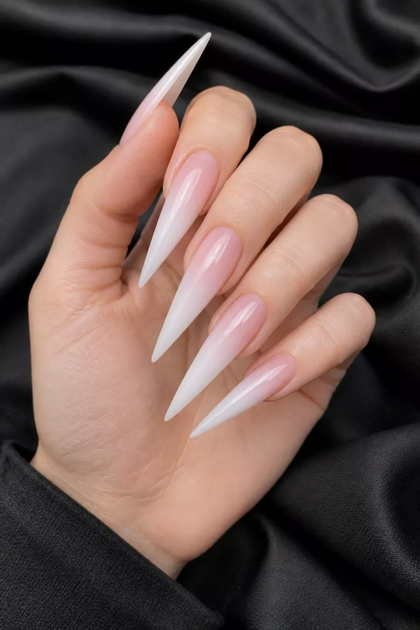 Milky White Natural Ombre close-up shot of a woman’s hand with five fingers showing extremely long, pointed stiletto nails painted a sheer milky white and pink ombré, appearing very natural and glossy, held over dark fabric.