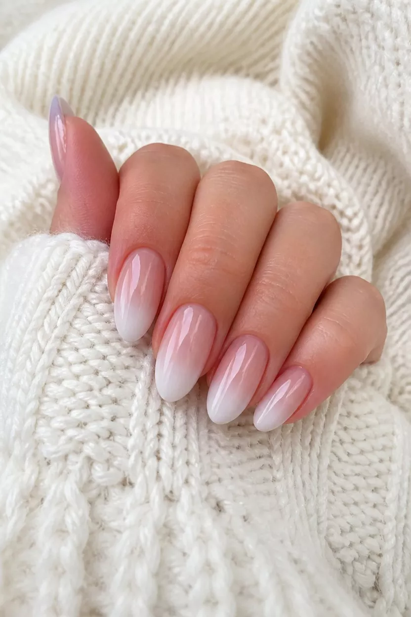 Milky Pink Ombre close-up shot of a woman’s hand with five fingers showing nails with medium-length, oval-to-almond nails showcasing a sheer, milky pink ombre, a white, cozy, textured sweater background.