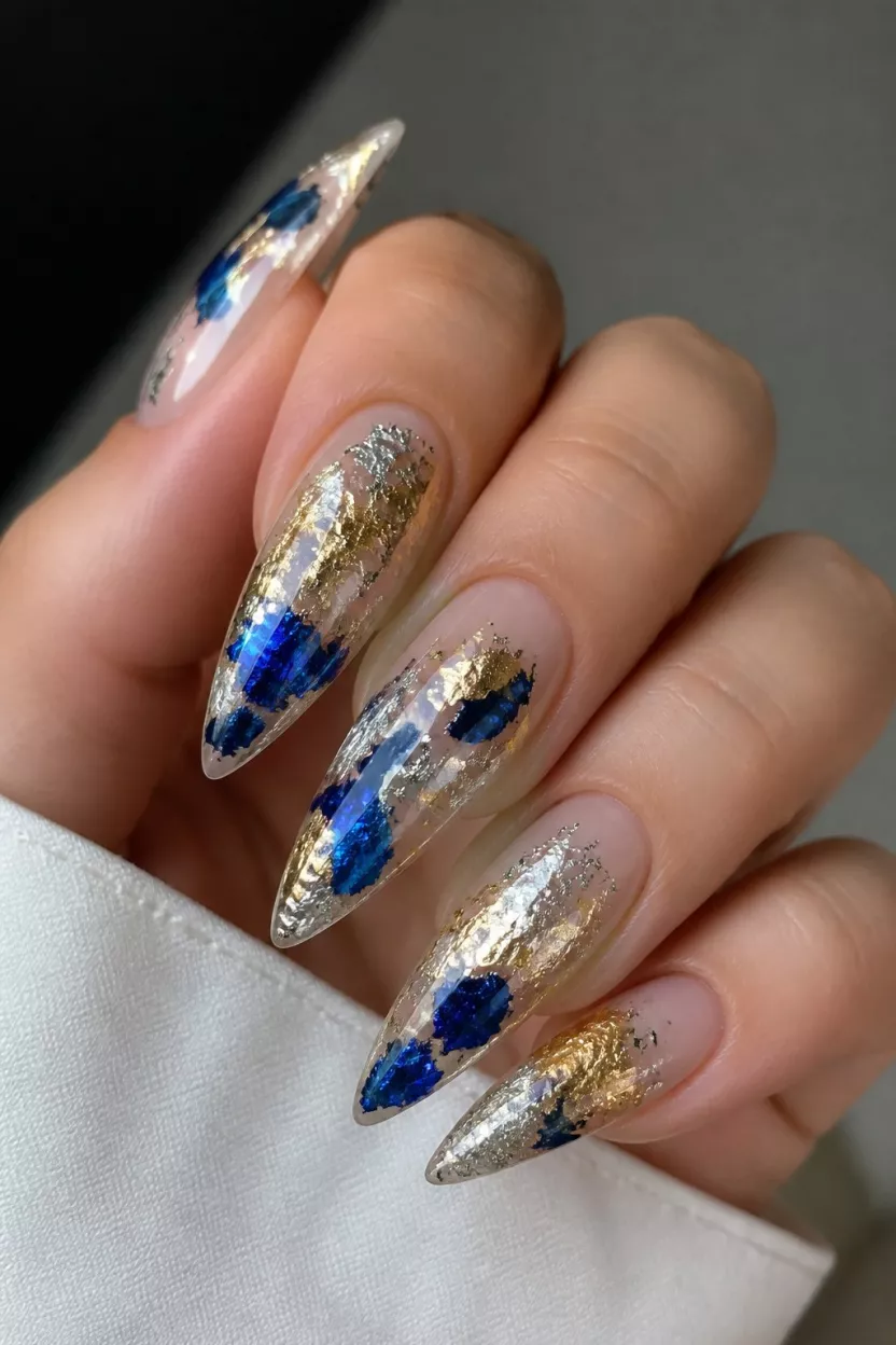 Celestial Shattered Glass close-up shot of a woman’s hand with five fingers showing nails with long, sharp stiletto nails designed with an abstract blue, silver, and gold foil overlay resembling shattered glass or celestial objects against a clear base.
