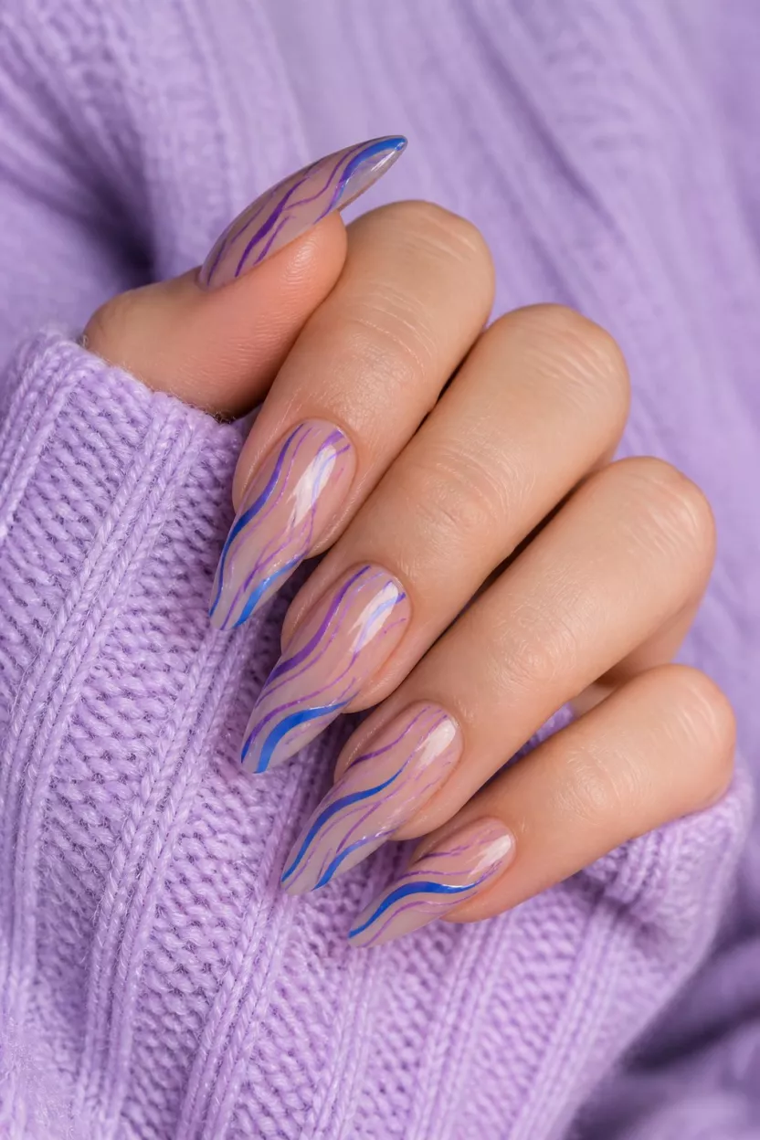 close-up shot of a woman’s hand with five fingers showing nails with long pointed nails have a sheer base with vibrant blue and purple curved lines, hand resting on a lavender sweater.