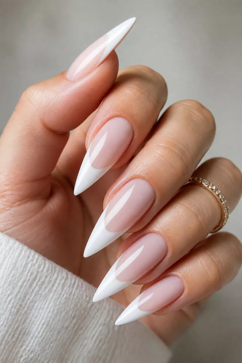 The Sharp White Stiletto close-up shot of a woman’s hand with five fingers showing nails with stiletto shaped nails show a translucent, milky pink base with elongated, sharp white French tips.