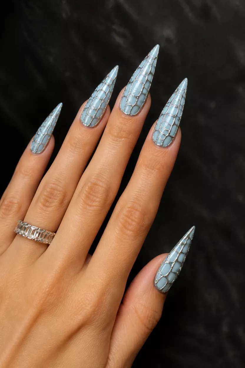 Blue Reptile Stiletto close-up shot of a woman’s hand with five fingers showing nails with stiletto nails adorned with intricate, elongated light blue designs resembling reptile or shell textures, accented with silver lines, against a dark textured background.