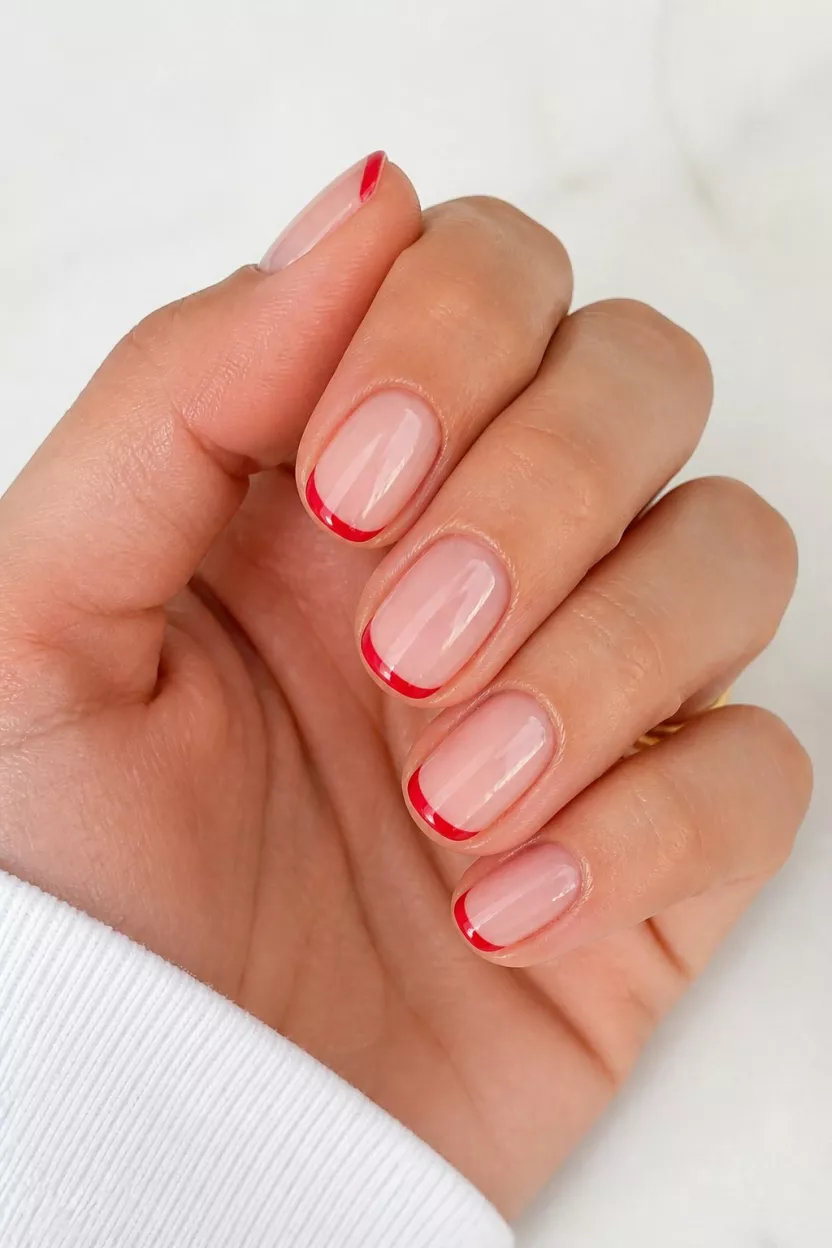 The Skinny Red French close-up shot of a woman’s hand with five fingers showing nails with short, rounded nails display a clean French tip design with a bright red border, set in a smooth, light background.