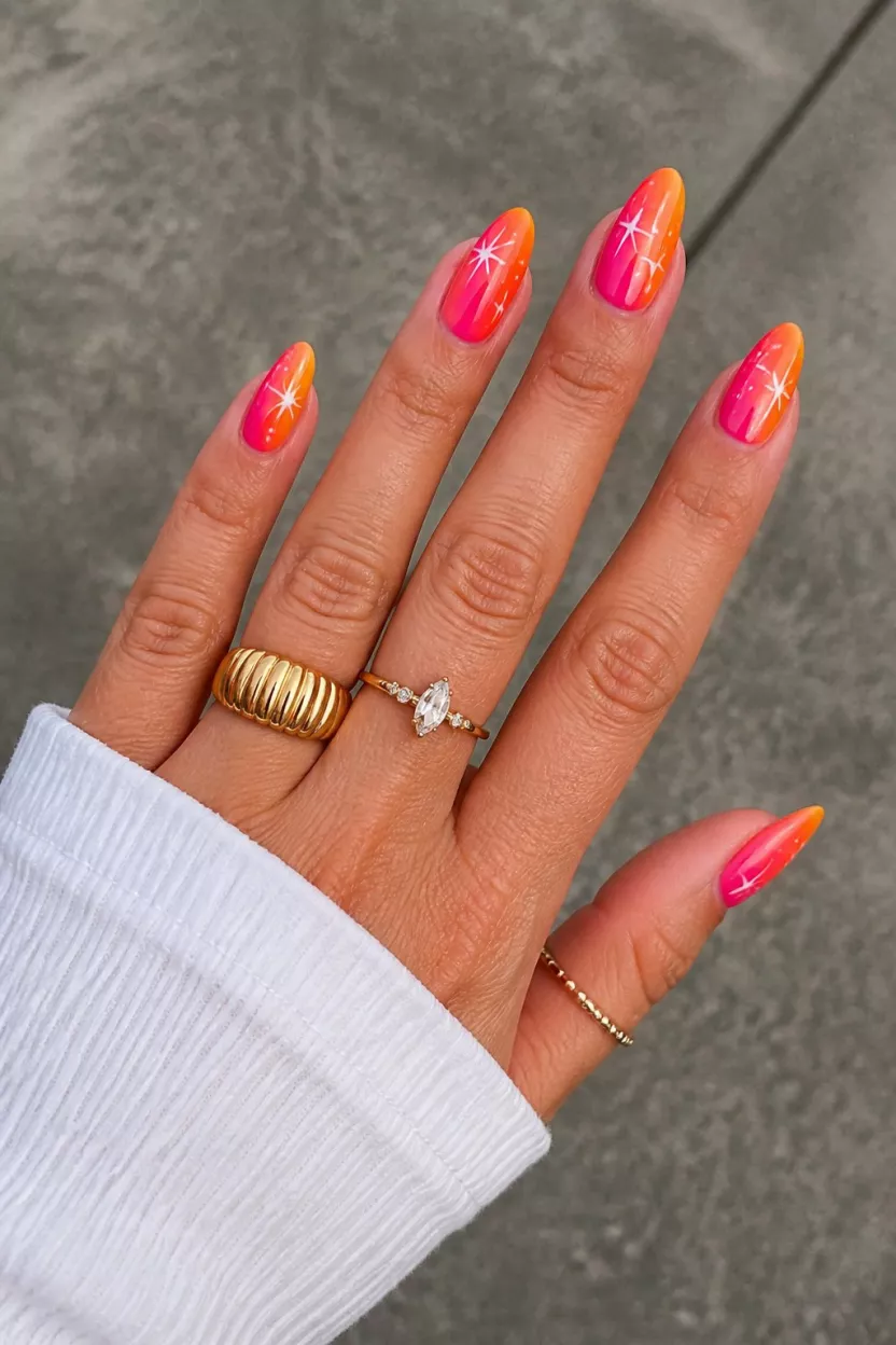 Sunset Starburst Gradient close-up shot of a woman’s hand with five fingers showing nails with almond shaped nails showing a vibrant pink to orange gradient with white star accents, accompanied by several gold-toned rings, on an outdoor gray surface.