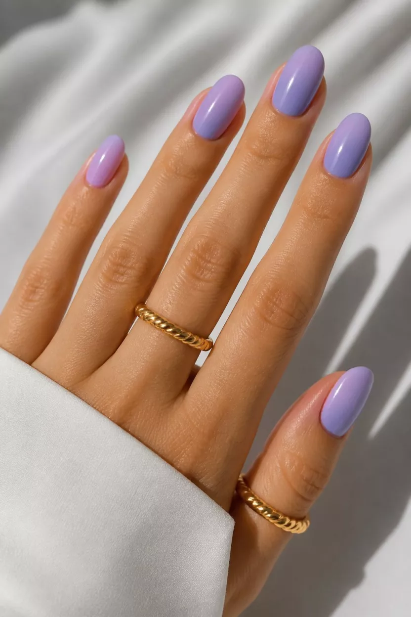 Lavender Fields close-up shot of a woman’s hand with five fingers showing nails with oval nails elegantly showcases a solid soft lavender polish, complemented by twisted gold toned rings.