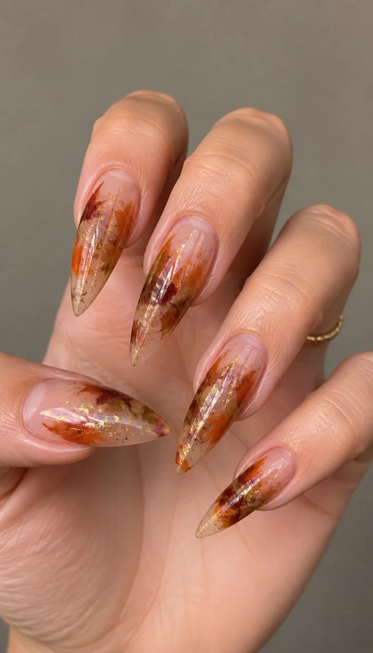 close-up shot of a woman’s hand with five fingers showing long, pointed nails featuring a sheer base with abstract autumnal color splashes and scattered glitter, neutral background.