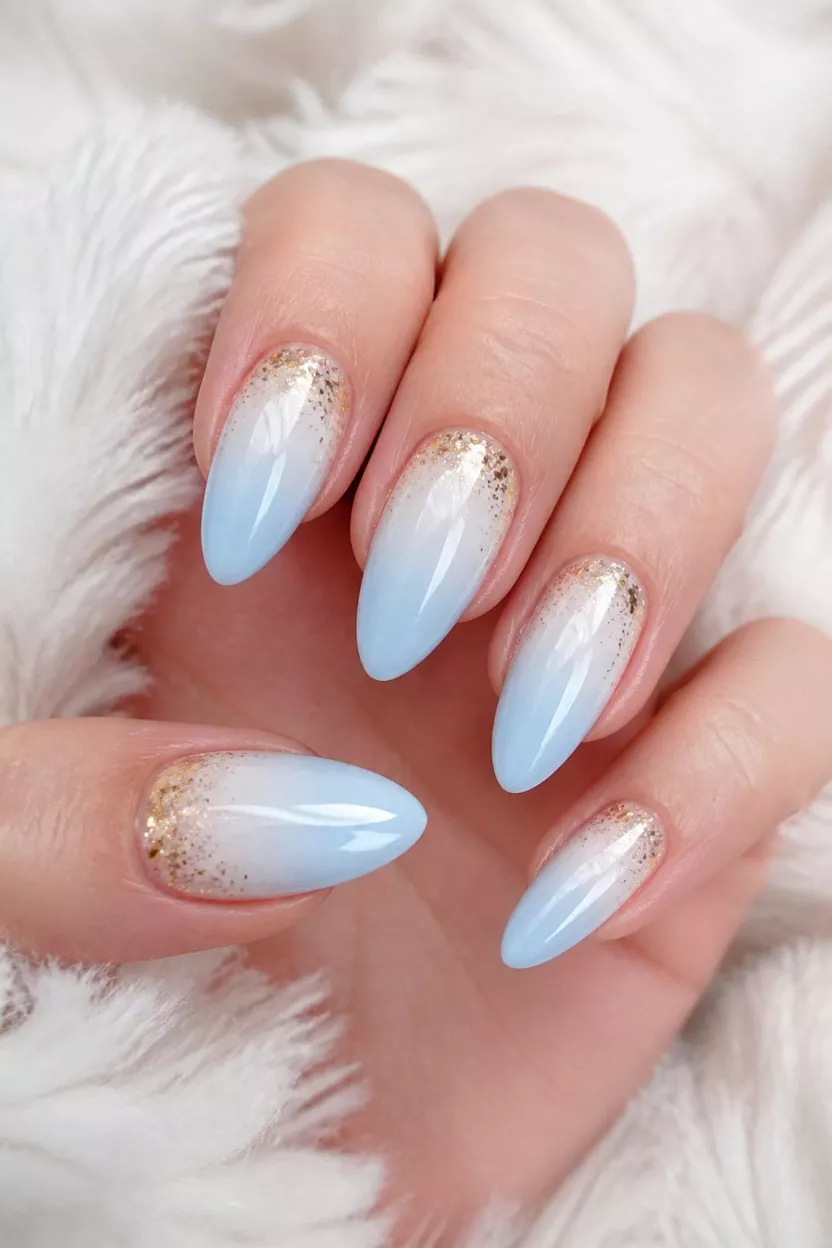 close-up shot of a woman’s hand with five fingers showing almond-shaped nails with an ombre design blending light blue milky white and gold glitter accents, soft fuzzy white material background