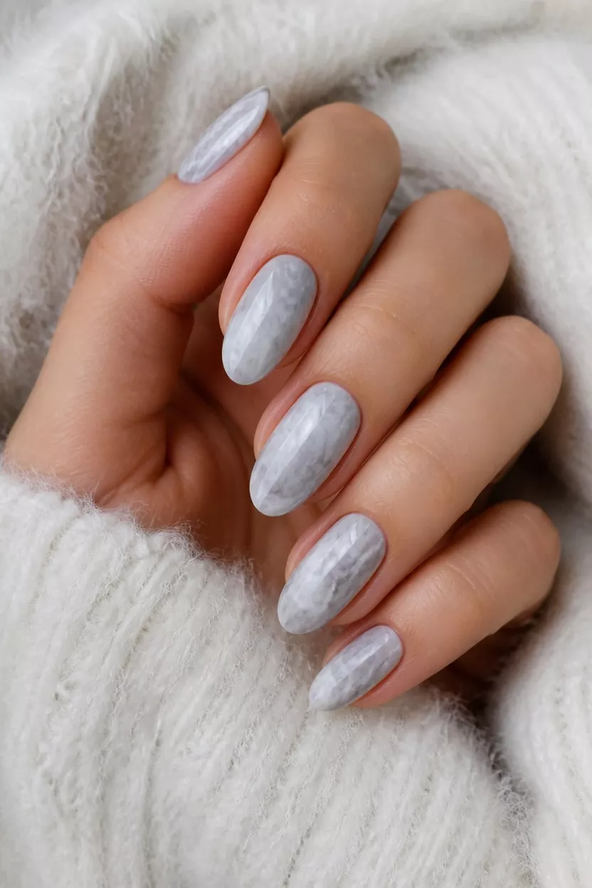 close-up shot of a woman’s hand with five fingers showing medium-length almond-shaped nails with a grey and white marbled or cloudy design, white fuzzy sweater background