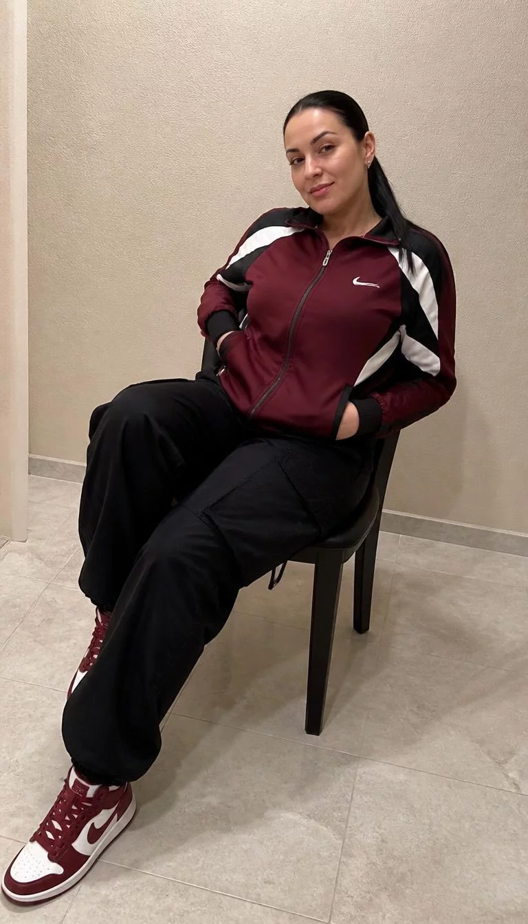 A photorealistic full body shot of a 33-year-old Eastern European woman with a round face, fair, broad nose, thin arched eyebrows, and thin lips. Her face features uneven eyebrows, visible pores, and subtle natural facial asymmetry. She has hooded brown eyes, pronounced cheekbones, closed-mouth smile, playful. She is curvy build with sleek black hair in a low ponytail and is wearing a black, white, and maroon track jacket, black cargo pants, and maroon and white sneakers, sitting on a chair, no people in the background, high-quality mobile photography, shot on an iPhone 14.
