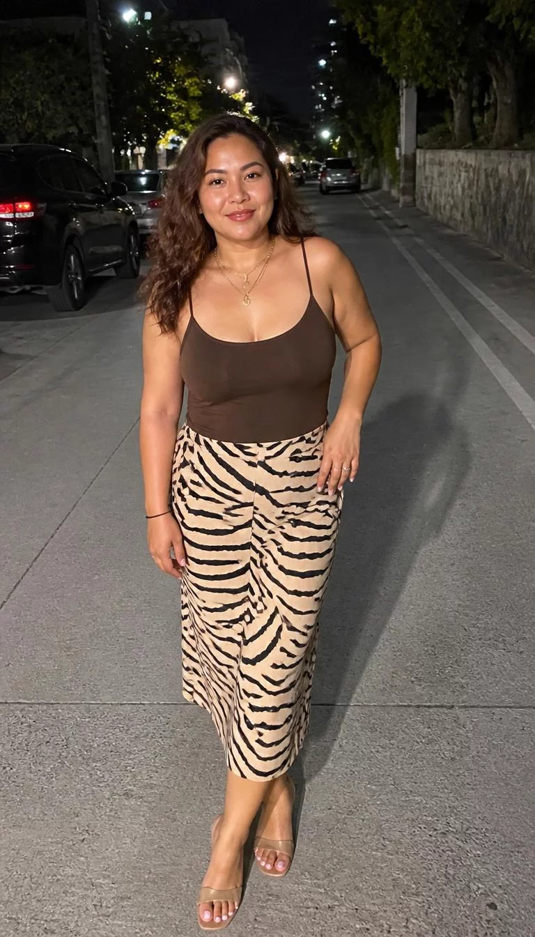 Brown Camisole And Tiger Print Midi Skirt A photorealistic full body shot of a 38-year-old Southeast Asian woman with a heart-shaped face, olive, broad nose, thick natural eyebrows, and full lips. Her face features beauty mark near the lip, faint under-eye shadows, and subtle natural facial asymmetry. She has upturned hazel eyes, rounded cheeks, broad smile, confident. She is average build with brown wavy hair and is wearing a brown camisole top, a beige tiger-print midi skirt, and layered gold necklaces, posing at night on an urban street with nearby parked cars, no people in the background, high-quality mobile photography, shot on an iPhone 14.