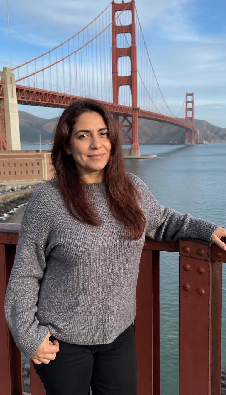 A photorealistic full body shot of a 47-year-old Middle Eastern woman with a oval face, deep brown, aquiline nose, thick natural eyebrows, and wide lips. Her face features dimples, a few laugh lines, and subtle natural facial asymmetry. She has almond-shaped hazel eyes, soft cheekbones, subtle smile, cheerful. She is average build with reddish-brown long hair styled loose hair and is wearing a grey sweater, posing near the orange Golden Gate Bridge span, everyday social media post aesthetic, high-quality mobile photography, shot on an iPhone 14.