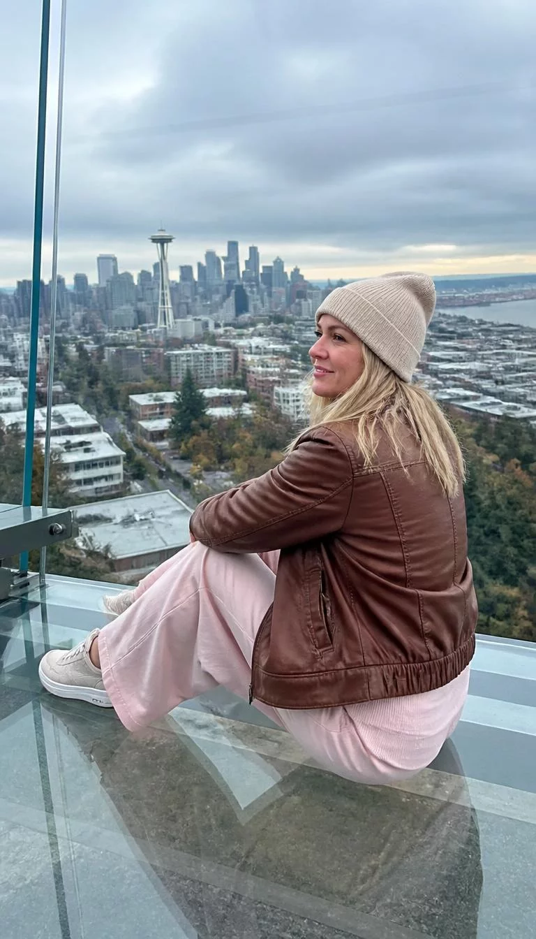 Beige Beanie And Brown Leather Jacket A photorealistic full body shot of a 39-year-old Northern European woman with a diamond face, fair, straight nose, feathered eyebrows, and thin lips. Her face features small scar on the eyebrow, a few laugh lines, and subtle natural facial asymmetry. She has upturned gray eyes, high cheekbones, closed-mouth smile, cheerful. She is average build with blonde hair hair and is wearing a beige beanie, brown leather jacket, and light pink wide-leg pants sits on a glass platform overlooking the Seattle cityscape, looking out at the view, no people in the background, high-quality mobile photography, shot on an iPhone 14.